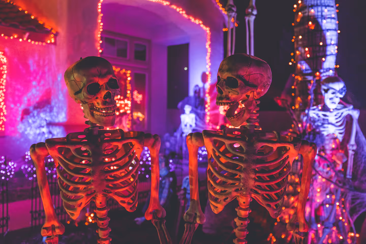 Dois esqueletos de plástico na frente de uma casa decorada com luzinhas laranja e roxas para o Halloween